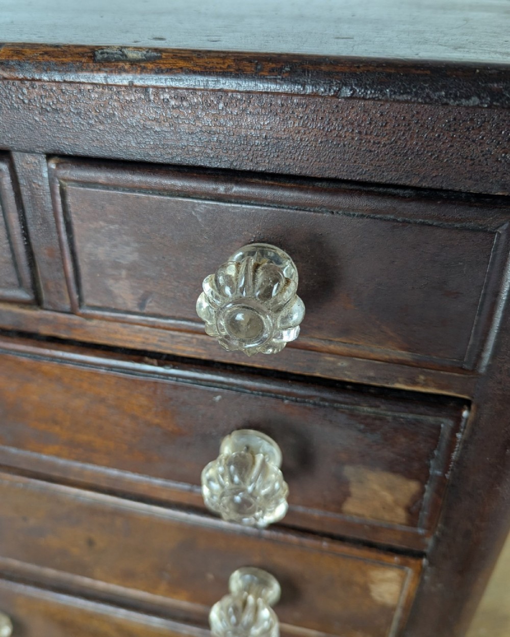 A dark brown Victorian miniature wooden chest of drawers with glass knobs and small turned feet. View XIII