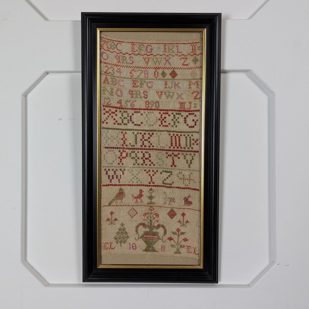 A vertical framed Georgian needlework sampler featuring rows of cross-stitched alphabets, numbers, and urn and bird motifs on a natural linen fabric, housed in a dark frame with a gilt inner slip.