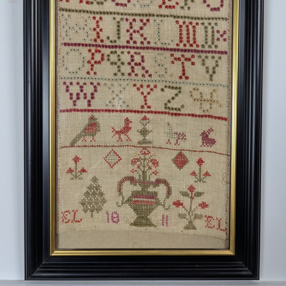 A vertical framed Georgian needlework sampler featuring rows of cross-stitched alphabets, numbers, and urn and bird motifs on a natural linen fabric, housed in a dark frame with a gilt inner slip. View III