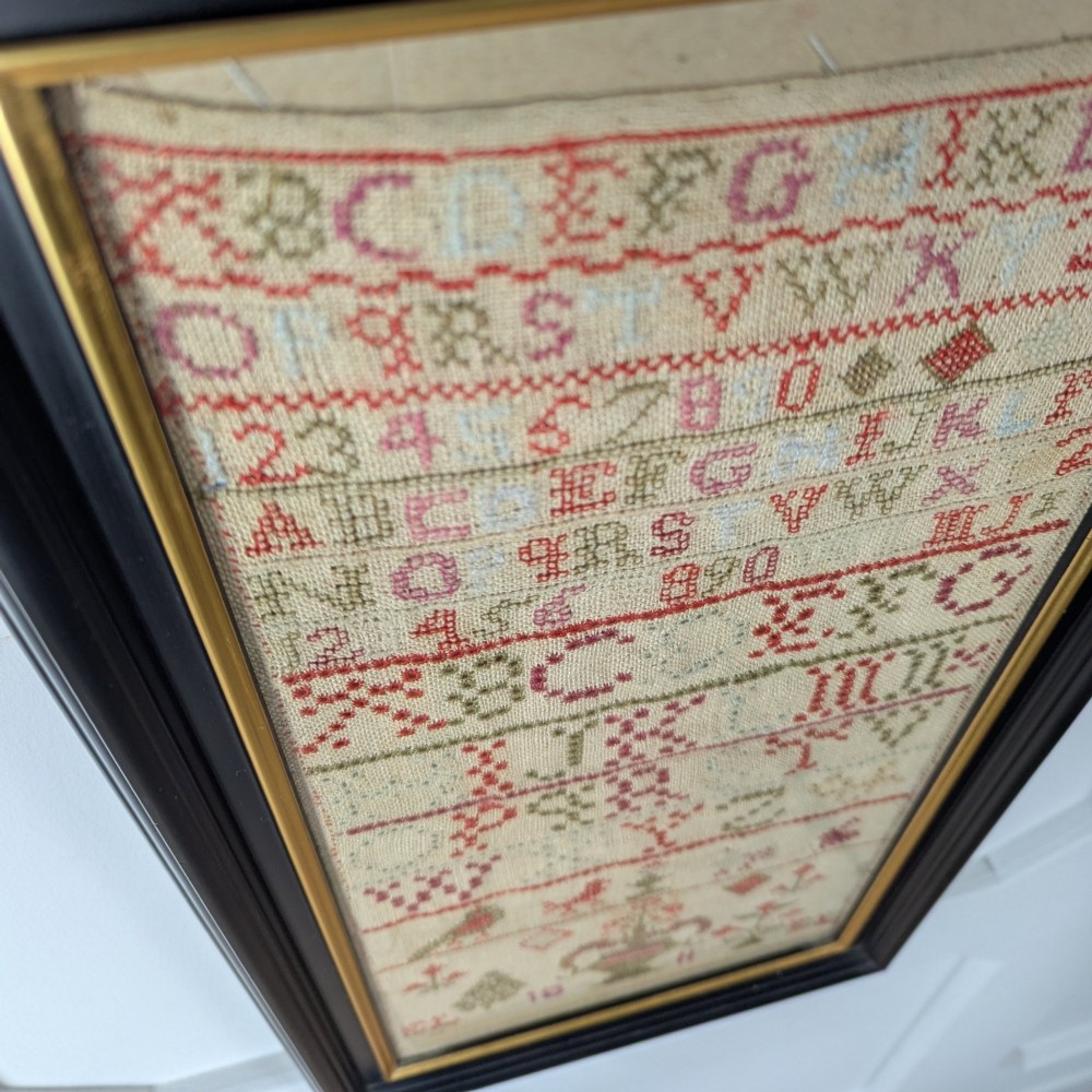 A vertical framed Georgian needlework sampler featuring rows of cross-stitched alphabets, numbers, and urn and bird motifs on a natural linen fabric, housed in a dark frame with a gilt inner slip. View IV
