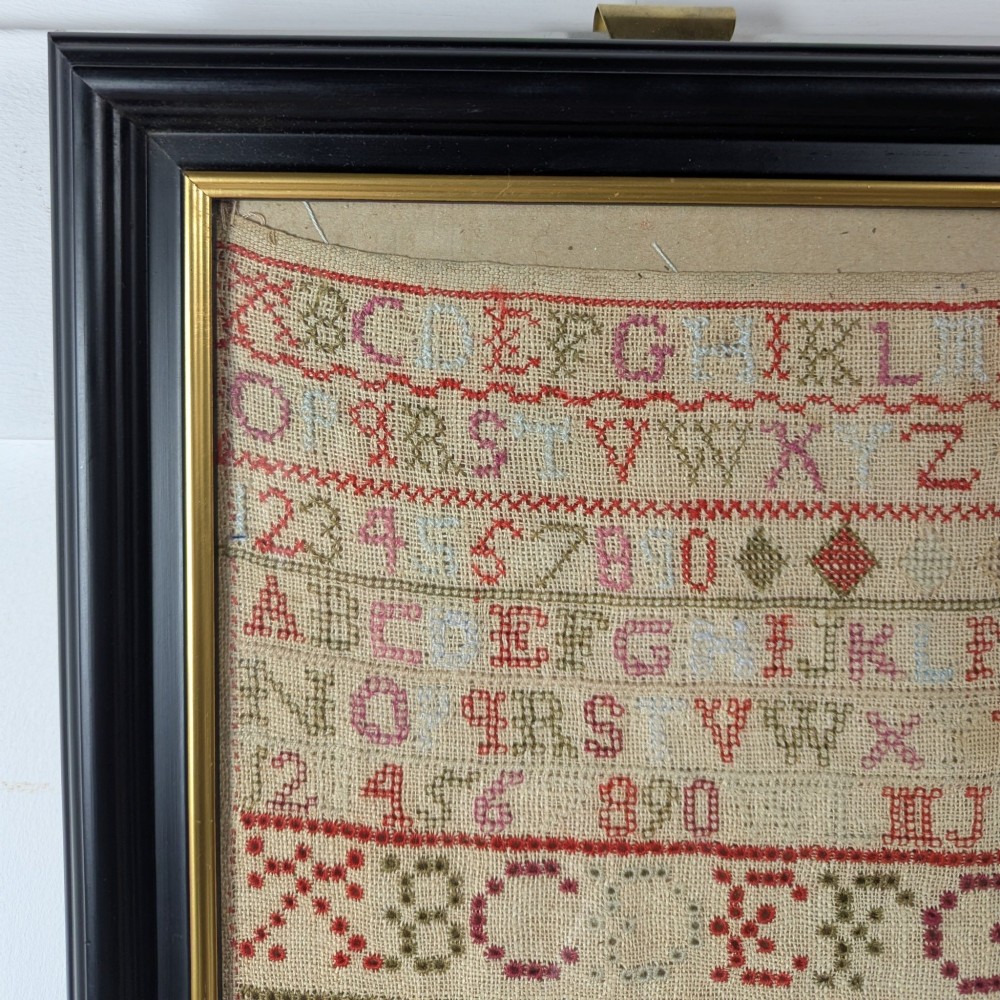 A vertical framed Georgian needlework sampler featuring rows of cross-stitched alphabets, numbers, and urn and bird motifs on a natural linen fabric, housed in a dark frame with a gilt inner slip. View V