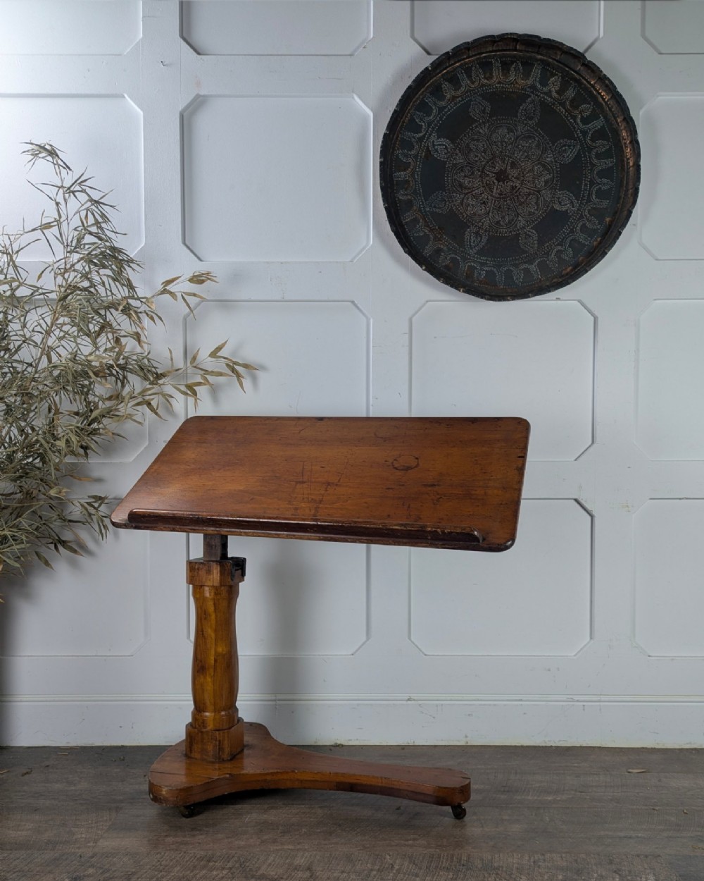 An early 20th-century pine reading table featuring an adjustable tilting top, a central turned support column, and a shaped base on small castors.