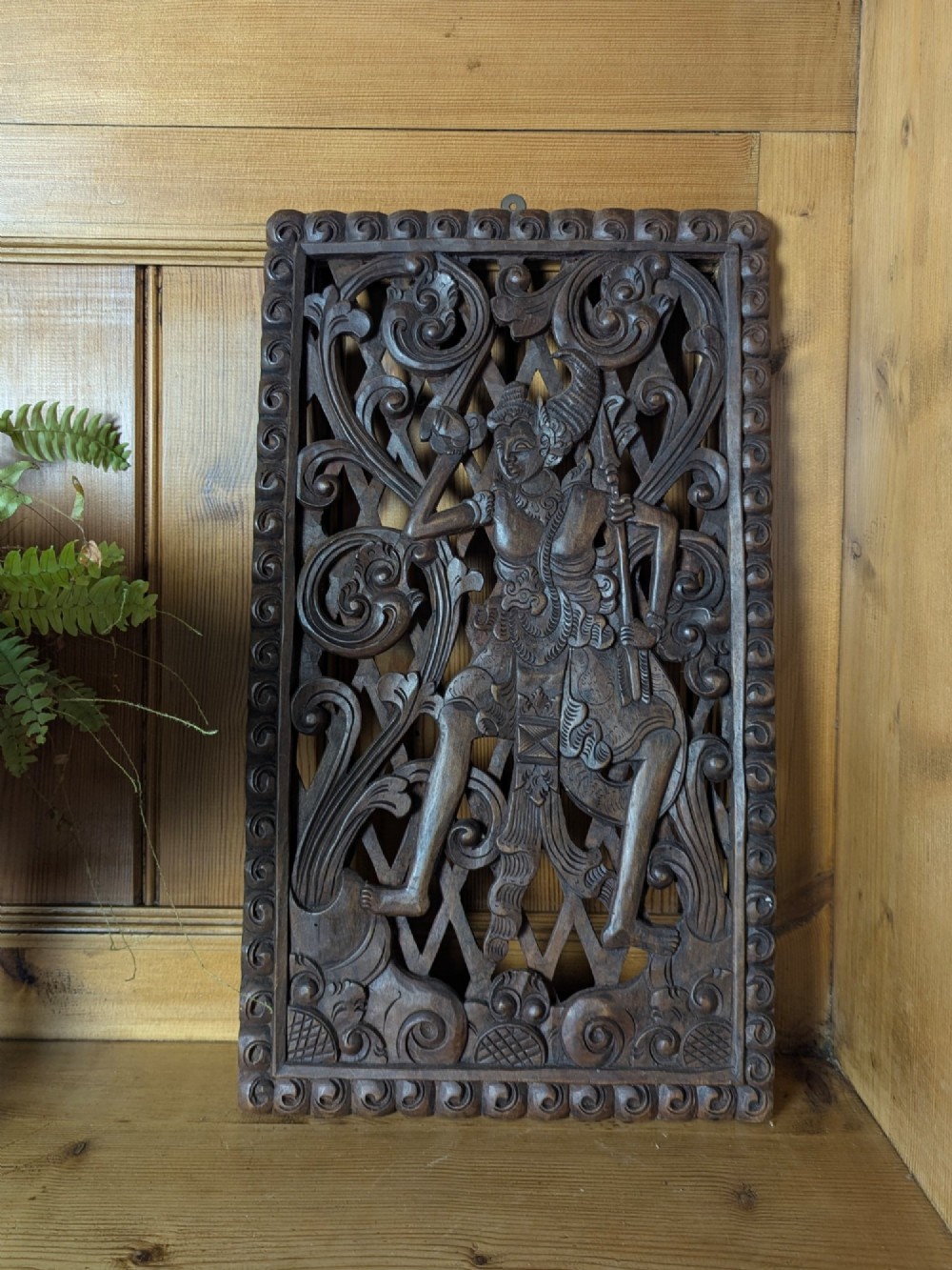 A rectangular dark teak wall panel hand-carved in high relief with an openwork Balinese mythological figure framed by intricate scrolling foliage.