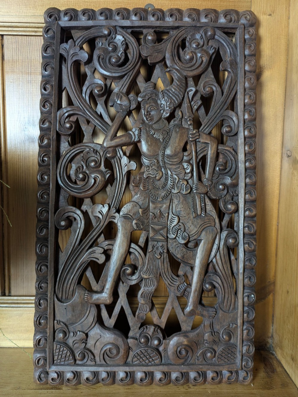 A rectangular dark teak wall panel hand-carved in high relief with an openwork Balinese mythological figure framed by intricate scrolling foliage. View II