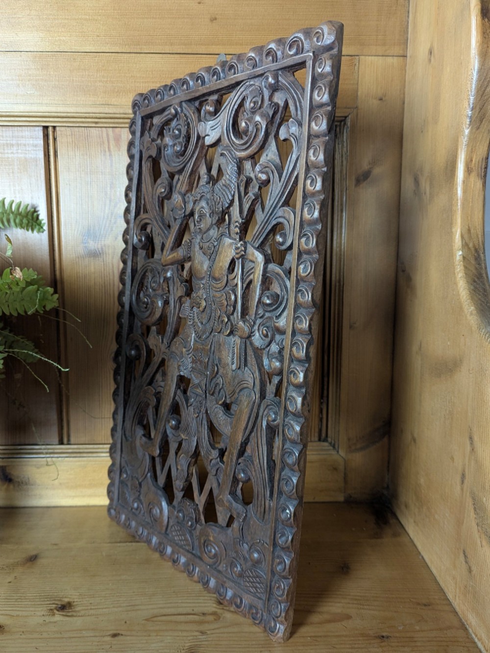 A rectangular dark teak wall panel hand-carved in high relief with an openwork Balinese mythological figure framed by intricate scrolling foliage. View V