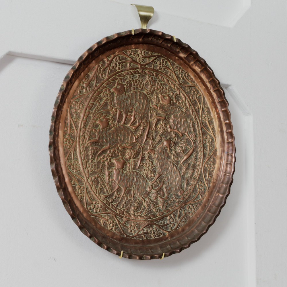 A circular hand-tooled copper charger tray featuring relief carvings of animals surrounded by foliage with a scalloped edge. View V