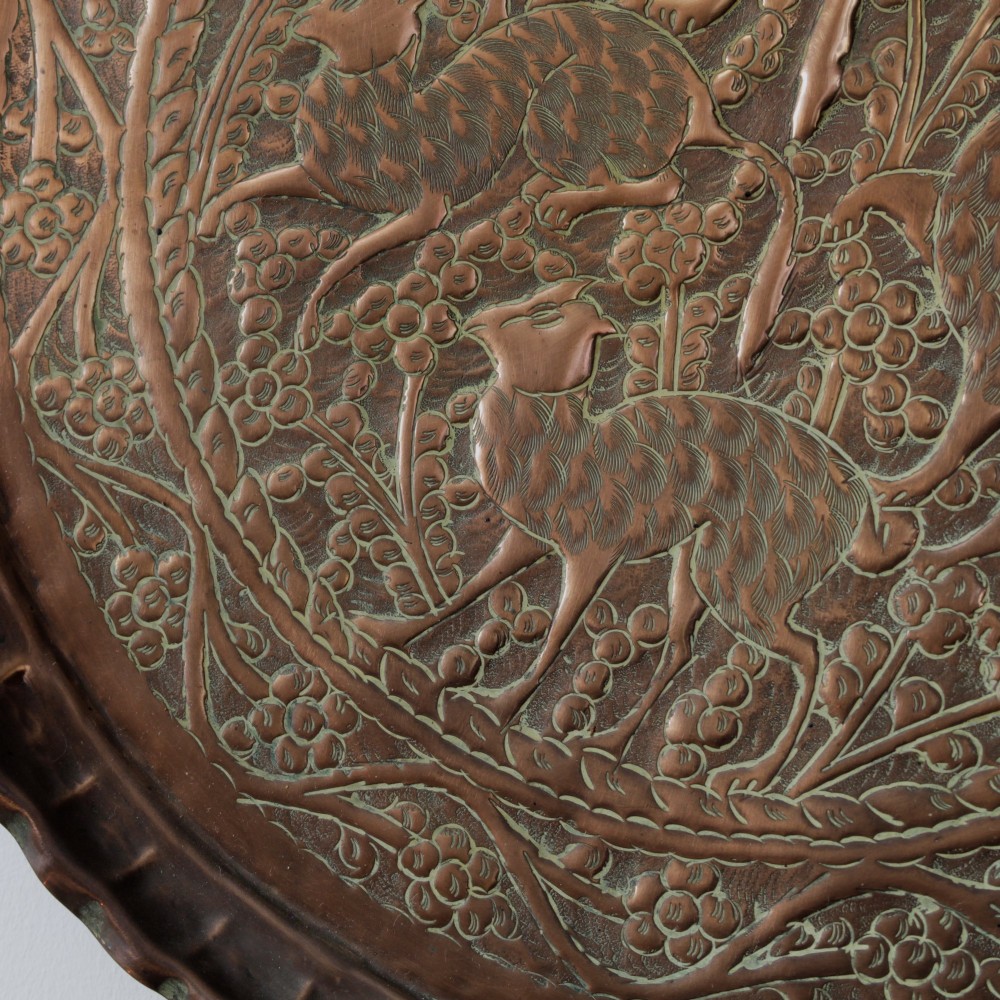 A circular hand-tooled copper charger tray featuring relief carvings of animals surrounded by foliage with a scalloped edge. View VI