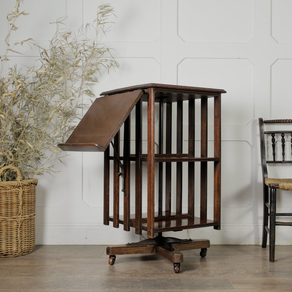 An Edwardian dark oak revolving bookcase featuring vertical slats, multiple shelf tiers, an adjustable folding reading rest, and a quadruped base on castors.