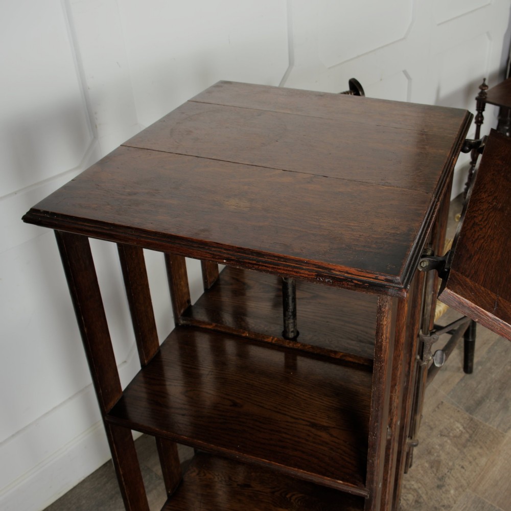 An Edwardian dark oak revolving bookcase featuring vertical slats, multiple shelf tiers, an adjustable folding reading rest, and a quadruped base on castors. View V