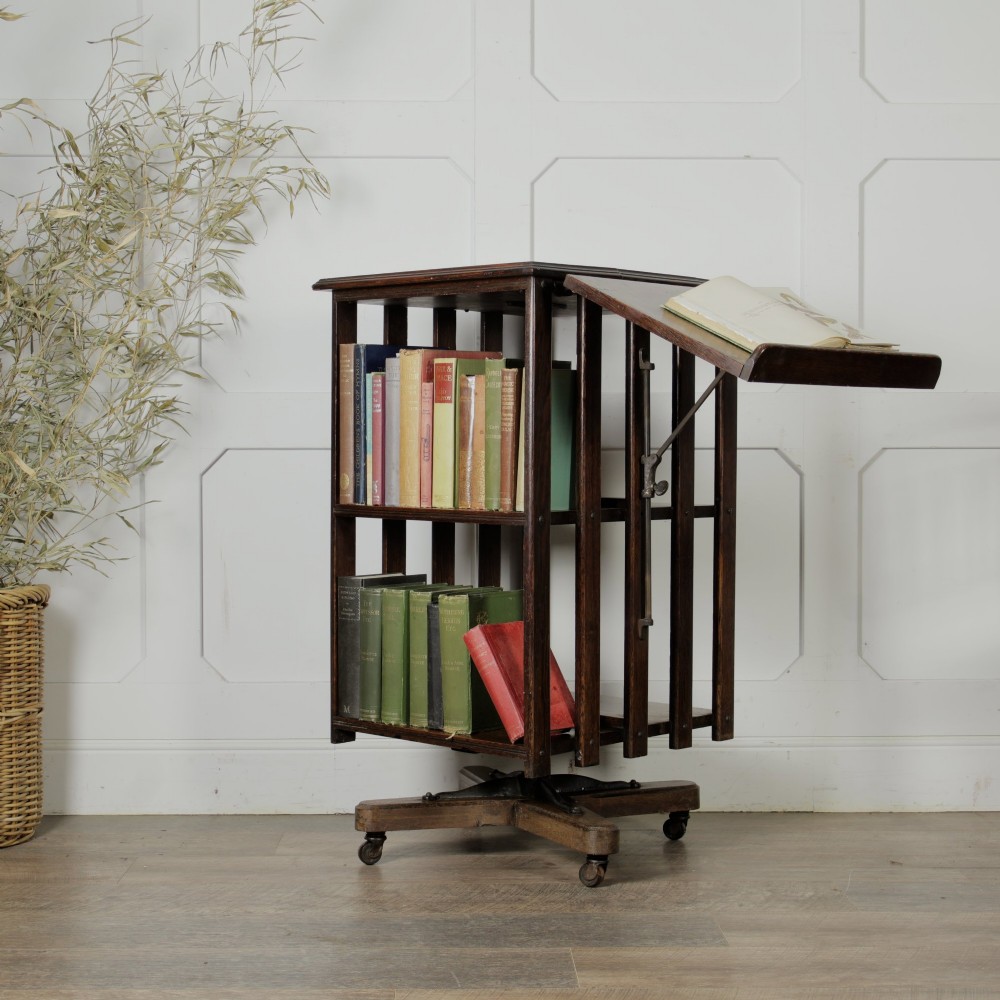 An Edwardian dark oak revolving bookcase featuring vertical slats, multiple shelf tiers, an adjustable folding reading rest, and a quadruped base on castors. View VI
