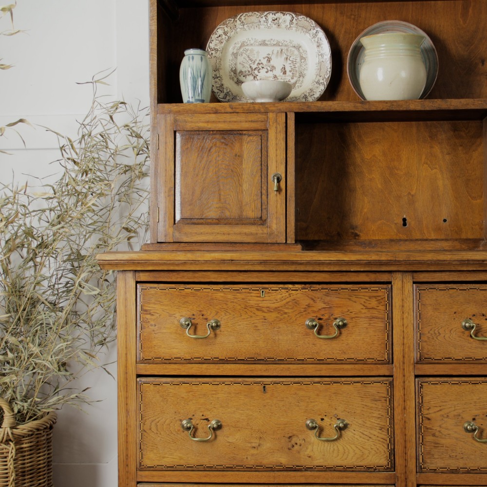A solid honey-coloured oak dresser featuring a top section with open shelving and small side cupboards, set upon a base with six drawers and brass swan-neck handles. View VI