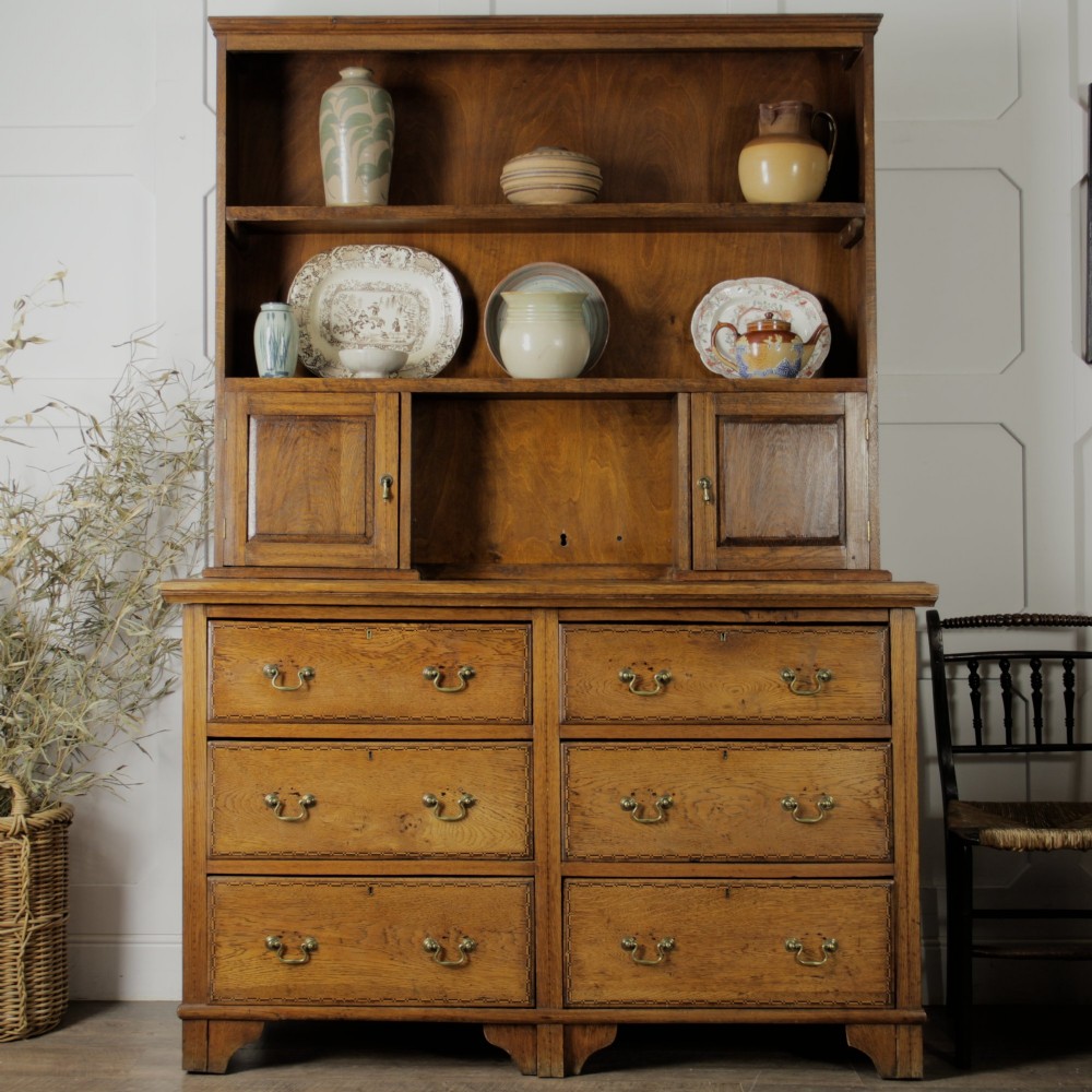 A solid honey-coloured oak dresser featuring a top section with open shelving and small side cupboards, set upon a base with six drawers and brass swan-neck handles. View VII