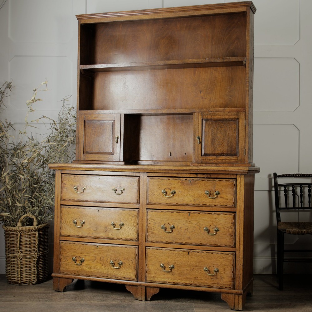 A solid honey-coloured oak dresser featuring a top section with open shelving and small side cupboards, set upon a base with six drawers and brass swan-neck handles.