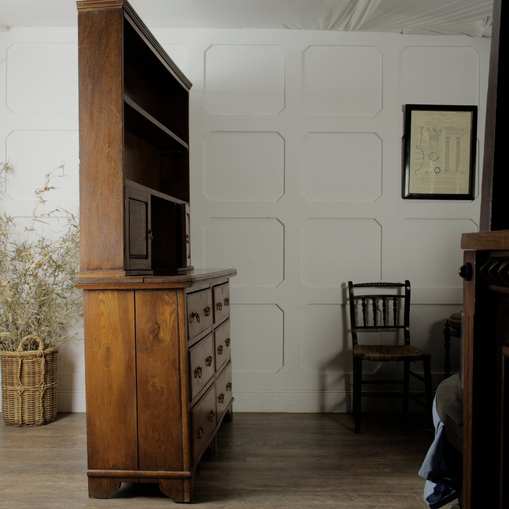 A solid honey-coloured oak dresser featuring a top section with open shelving and small side cupboards, set upon a base with six drawers and brass swan-neck handles. View IX