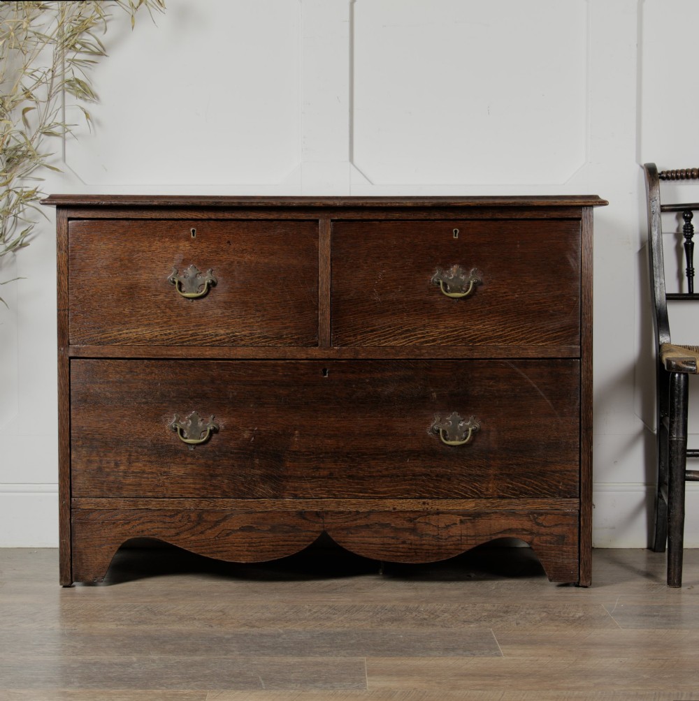 A dark solid oak chest of drawers featuring two upper drawers over a wide lower drawer with brass swan-neck handles and a shaped bottom apron. View II