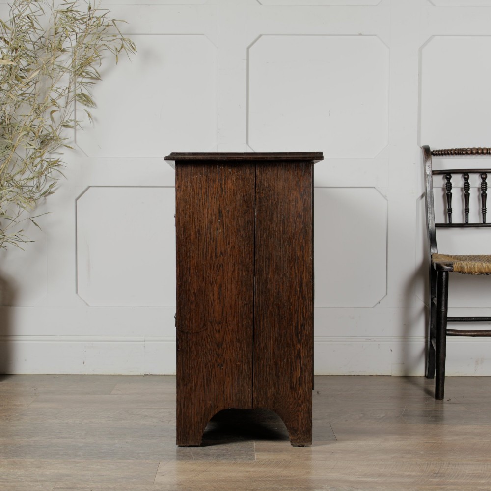 A dark solid oak chest of drawers featuring two upper drawers over a wide lower drawer with brass swan-neck handles and a shaped bottom apron. View III