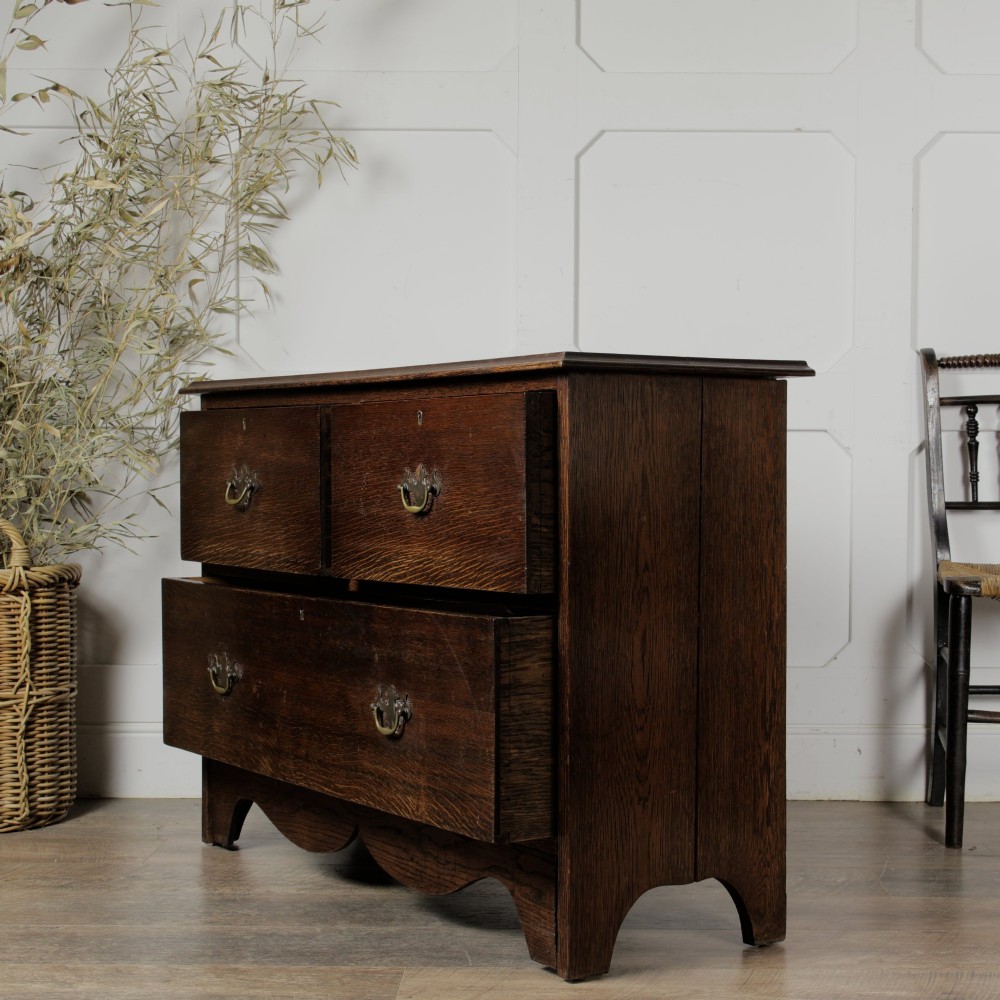 A dark solid oak chest of drawers featuring two upper drawers over a wide lower drawer with brass swan-neck handles and a shaped bottom apron. View IV