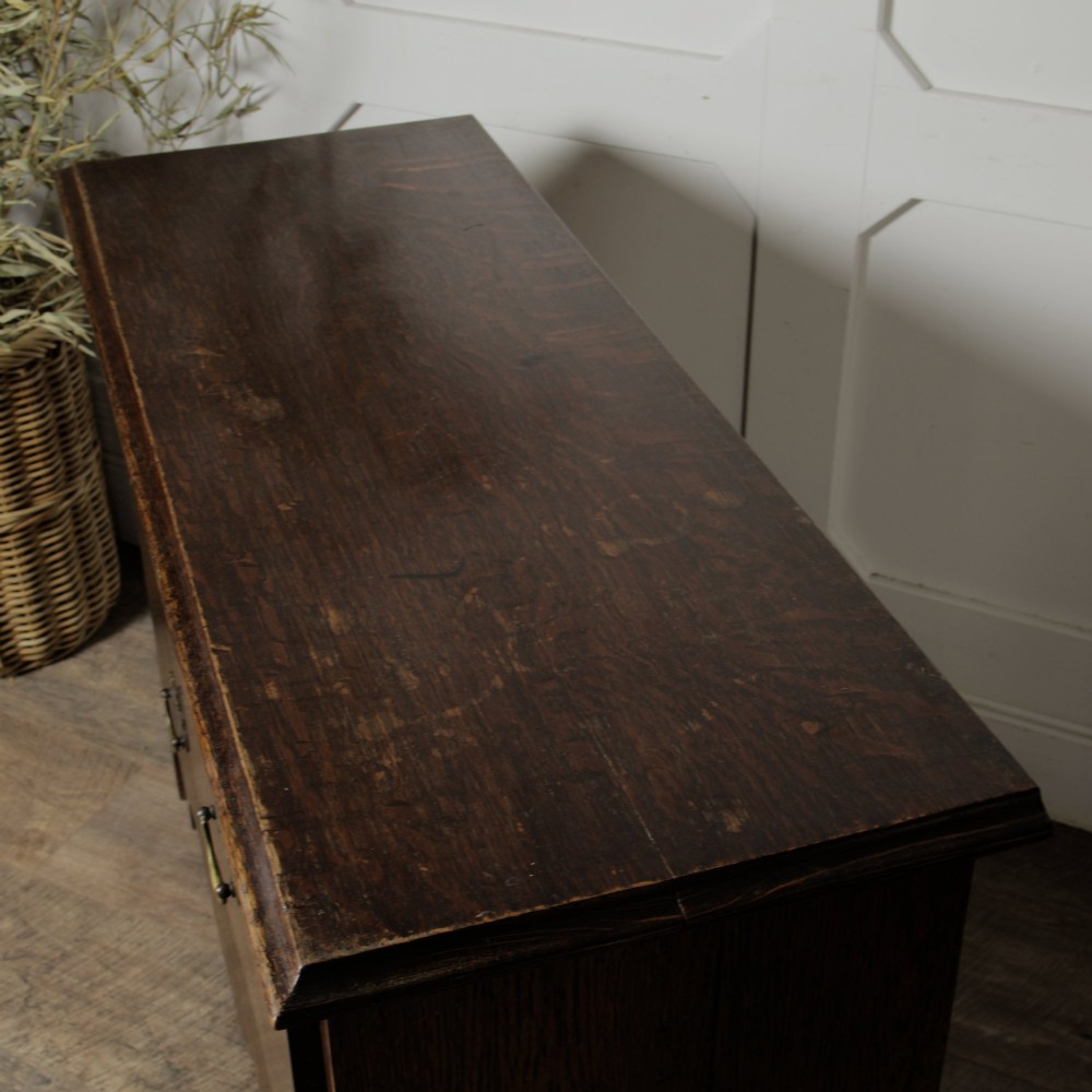 A dark solid oak chest of drawers featuring two upper drawers over a wide lower drawer with brass swan-neck handles and a shaped bottom apron. View V