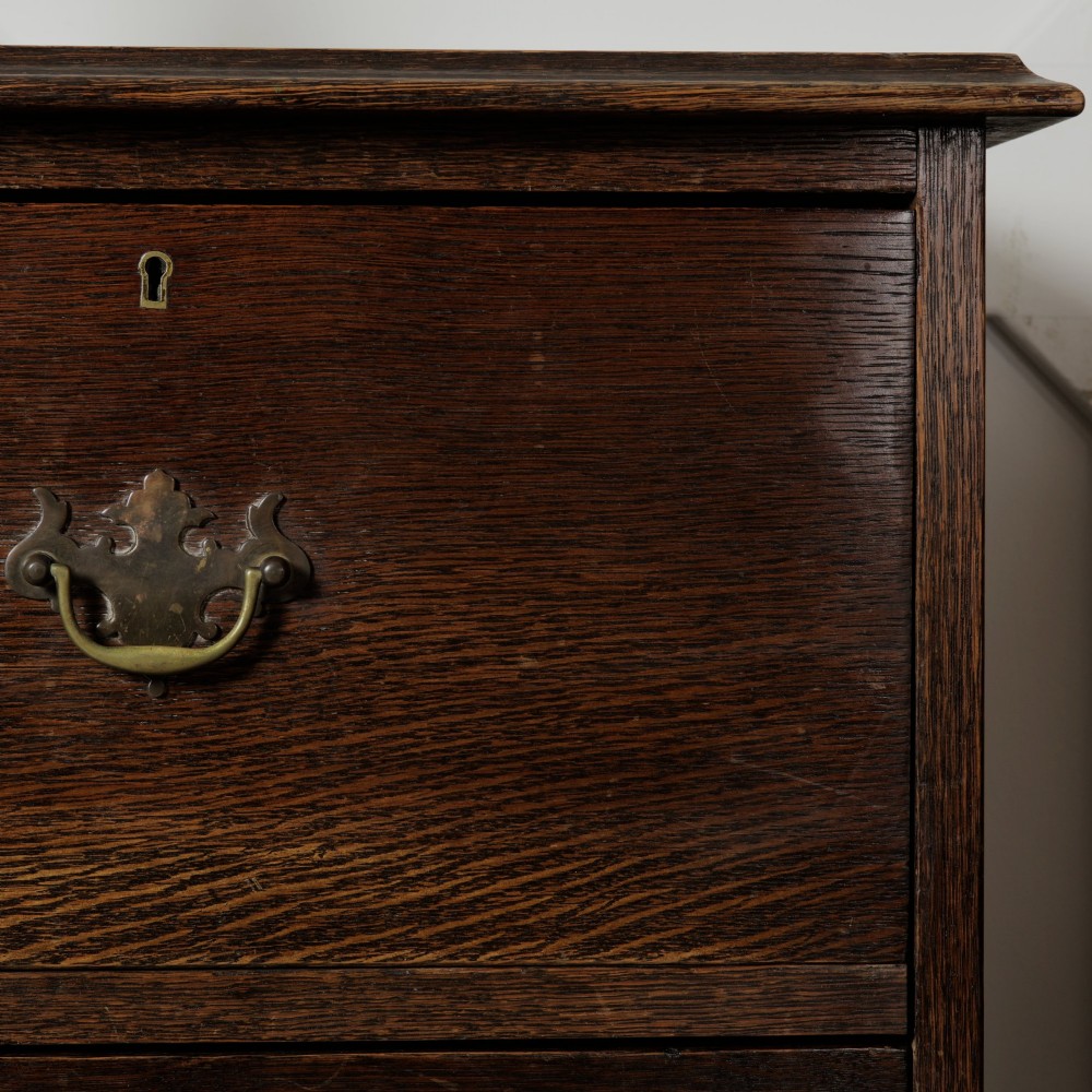A dark solid oak chest of drawers featuring two upper drawers over a wide lower drawer with brass swan-neck handles and a shaped bottom apron. View VII