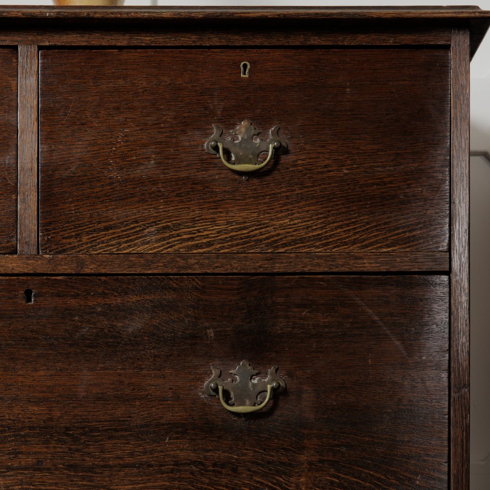 A dark solid oak chest of drawers featuring two upper drawers over a wide lower drawer with brass swan-neck handles and a shaped bottom apron. View VIII