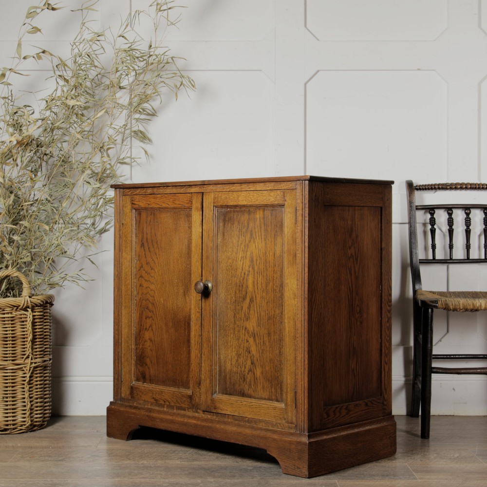 A solid oak low cupboard featuring two panelled doors with a central wooden knob handle set on a shaped plinth base. View II