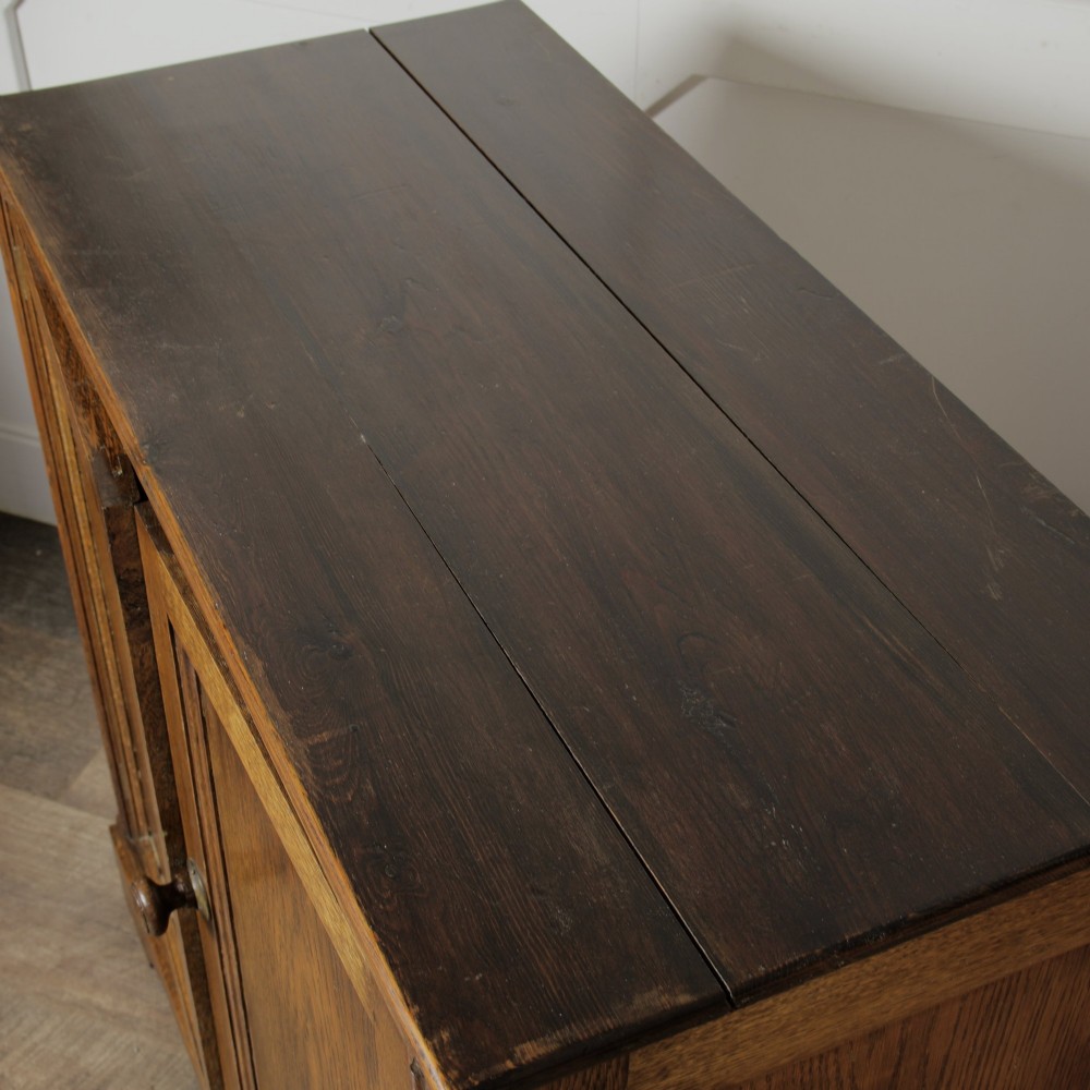 A solid oak low cupboard featuring two panelled doors with a central wooden knob handle set on a shaped plinth base. View VII