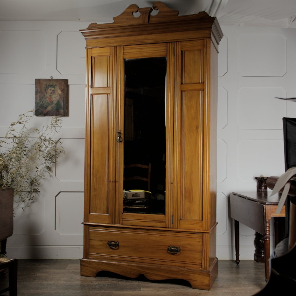 An early 20th-century satinwood wardrobe featuring a central mirrored door, carved top pediment, panelled sides, and a single lower drawer with metal drop handles.