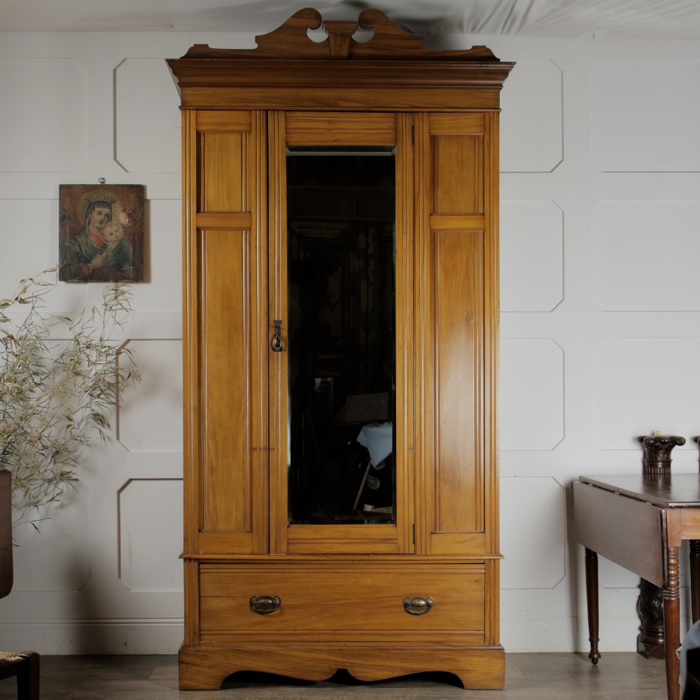 An early 20th-century satinwood wardrobe featuring a central mirrored door, carved top pediment, panelled sides, and a single lower drawer with metal drop handles. View III