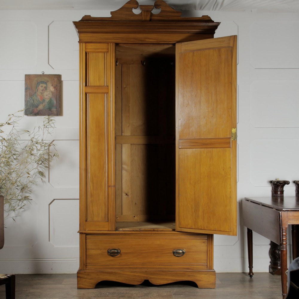 An early 20th-century satinwood wardrobe featuring a central mirrored door, carved top pediment, panelled sides, and a single lower drawer with metal drop handles. View IV