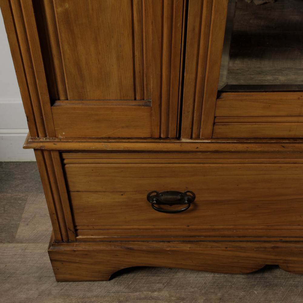 An early 20th-century satinwood wardrobe featuring a central mirrored door, carved top pediment, panelled sides, and a single lower drawer with metal drop handles. View VII