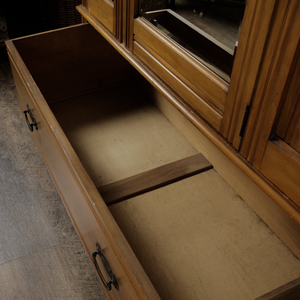An early 20th-century satinwood wardrobe featuring a central mirrored door, carved top pediment, panelled sides, and a single lower drawer with metal drop handles. View VIII