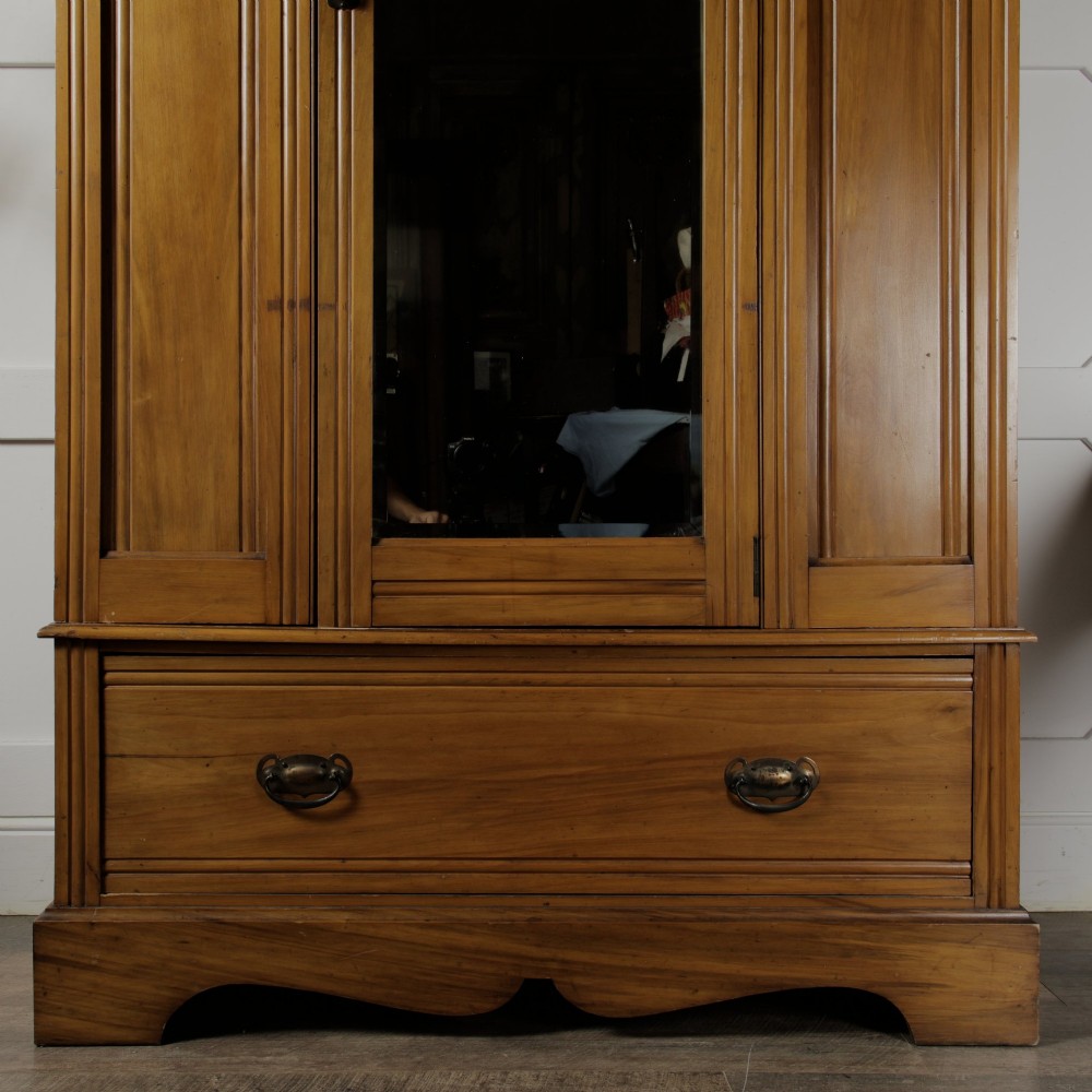 An early 20th-century satinwood wardrobe featuring a central mirrored door, carved top pediment, panelled sides, and a single lower drawer with metal drop handles. View IX