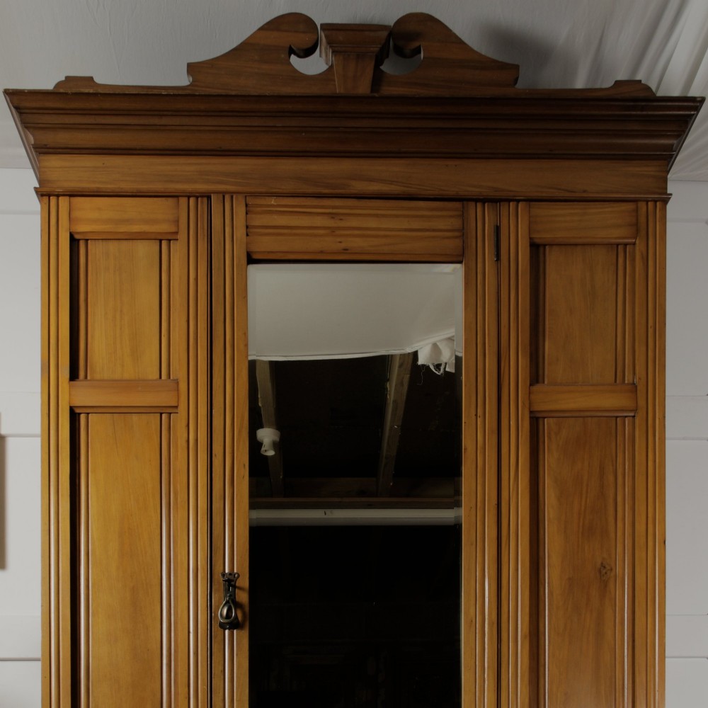 An early 20th-century satinwood wardrobe featuring a central mirrored door, carved top pediment, panelled sides, and a single lower drawer with metal drop handles. View X