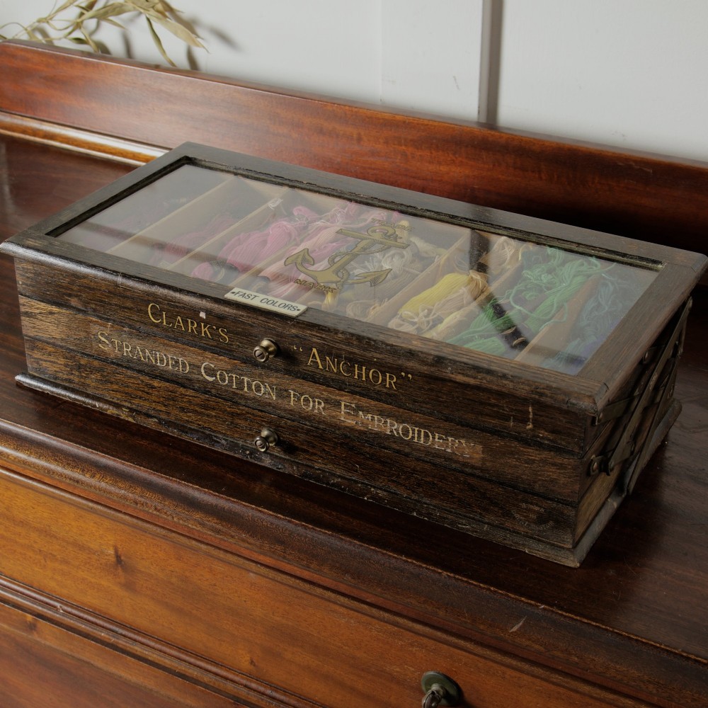 A dark wooden multi-tiered cantilever haberdashery display box with a glass lid bearing a yellow anchor logo, shown open with thread spools inside. View V