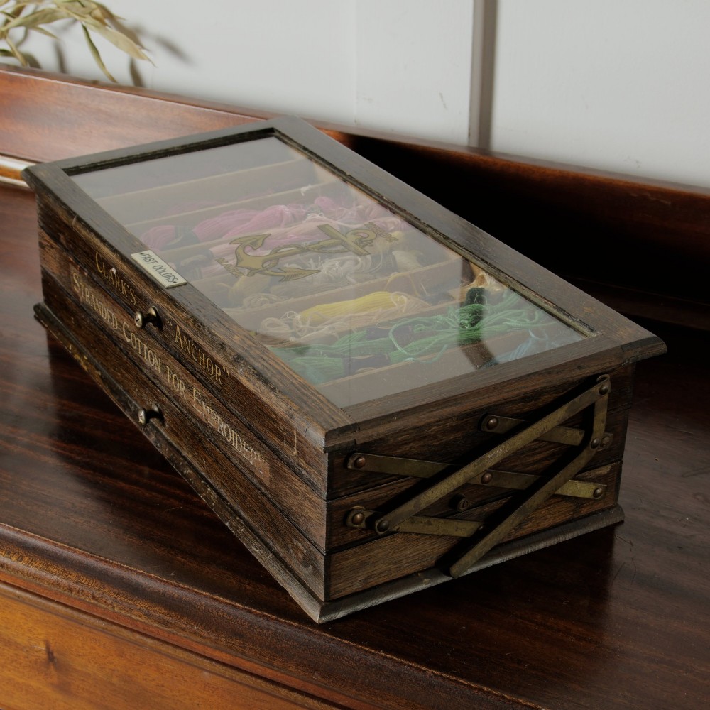 A dark wooden multi-tiered cantilever haberdashery display box with a glass lid bearing a yellow anchor logo, shown open with thread spools inside. View VI