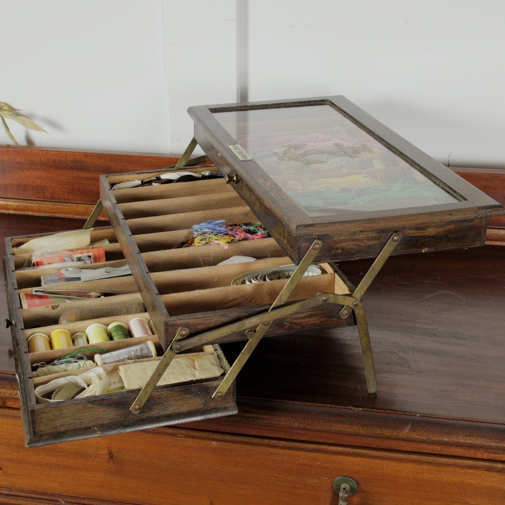 A dark wooden multi-tiered cantilever haberdashery display box with a glass lid bearing a yellow anchor logo, shown open with thread spools inside. View VII