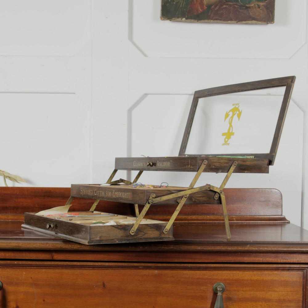 A dark wooden multi-tiered cantilever haberdashery display box with a glass lid bearing a yellow anchor logo, shown open with thread spools inside. View IX