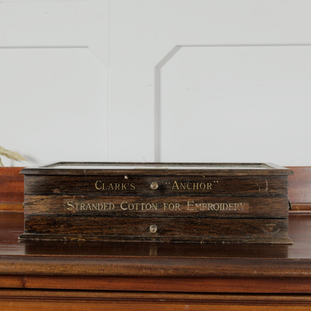 A dark wooden multi-tiered cantilever haberdashery display box with a glass lid bearing a yellow anchor logo, shown open with thread spools inside. View X