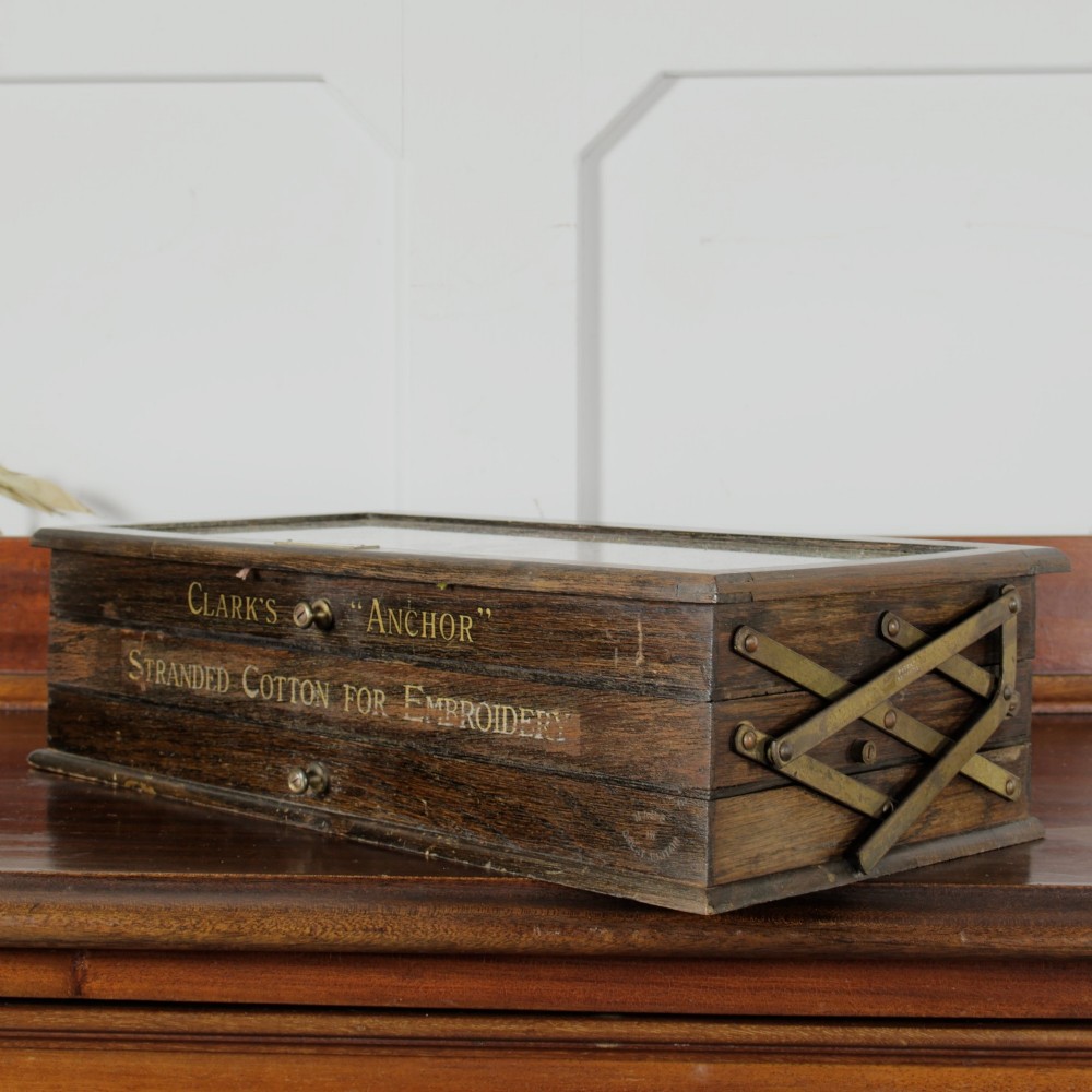A dark wooden multi-tiered cantilever haberdashery display box with a glass lid bearing a yellow anchor logo, shown open with thread spools inside. View XIV