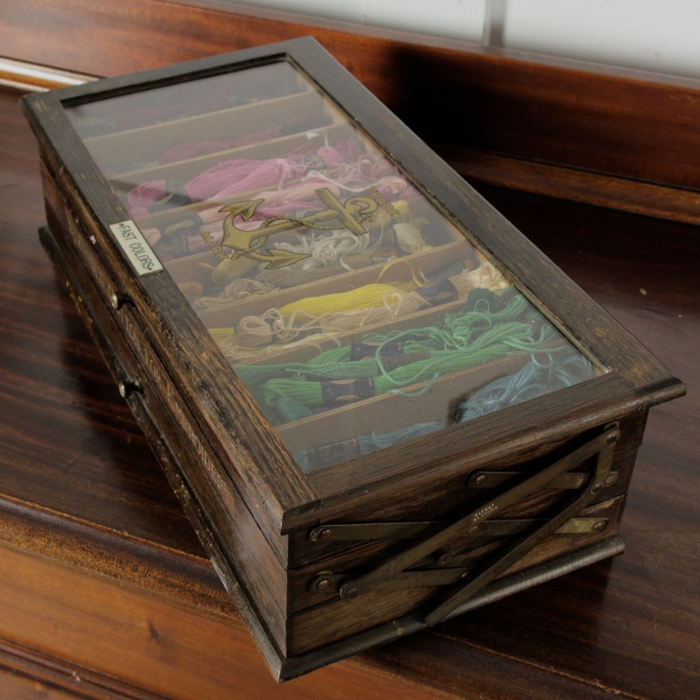 A dark wooden multi-tiered cantilever haberdashery display box with a glass lid bearing a yellow anchor logo, shown open with thread spools inside. View XV