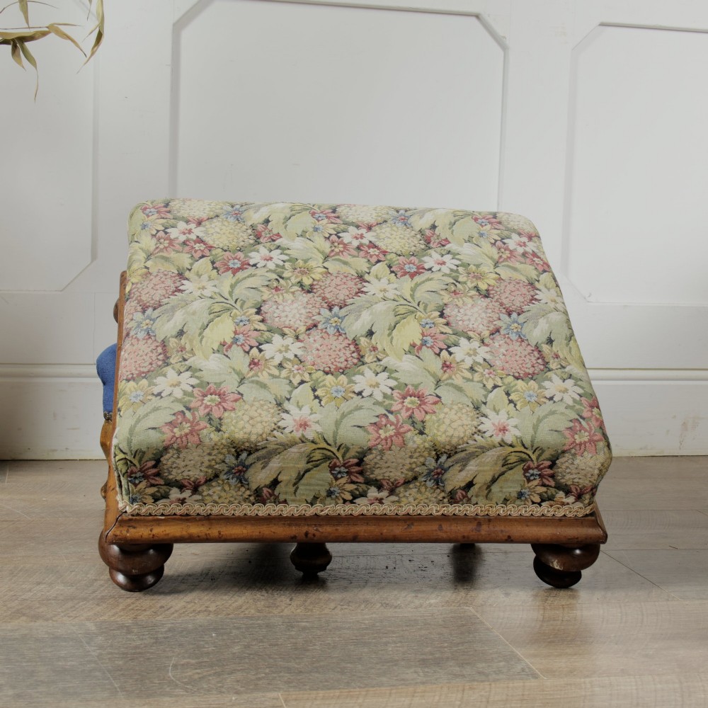 A low Victorian square footstool with green floral tapestry upholstery, decorative braid trim, and turned mahogany feet. View III
