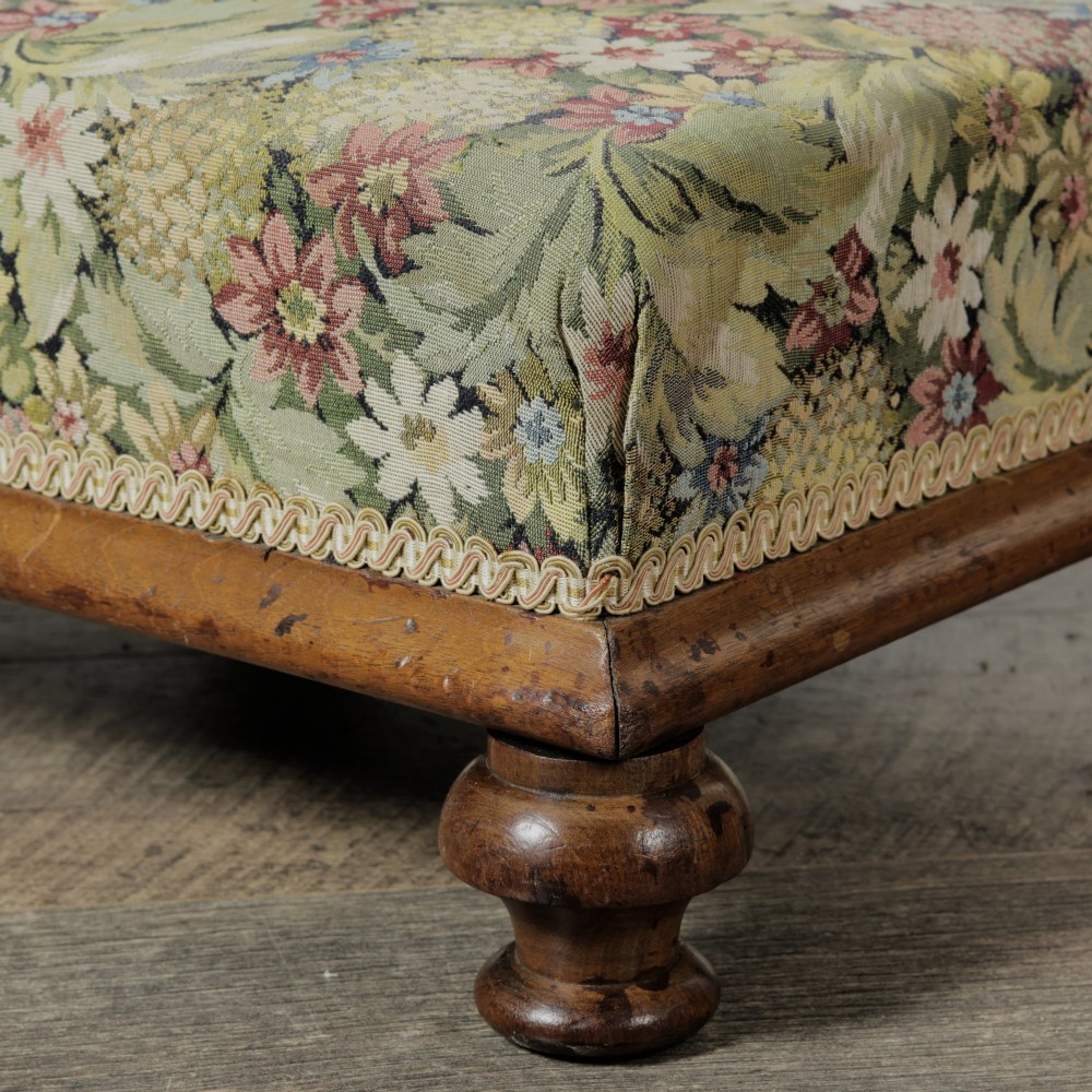 A low Victorian square footstool with green floral tapestry upholstery, decorative braid trim, and turned mahogany feet. View V