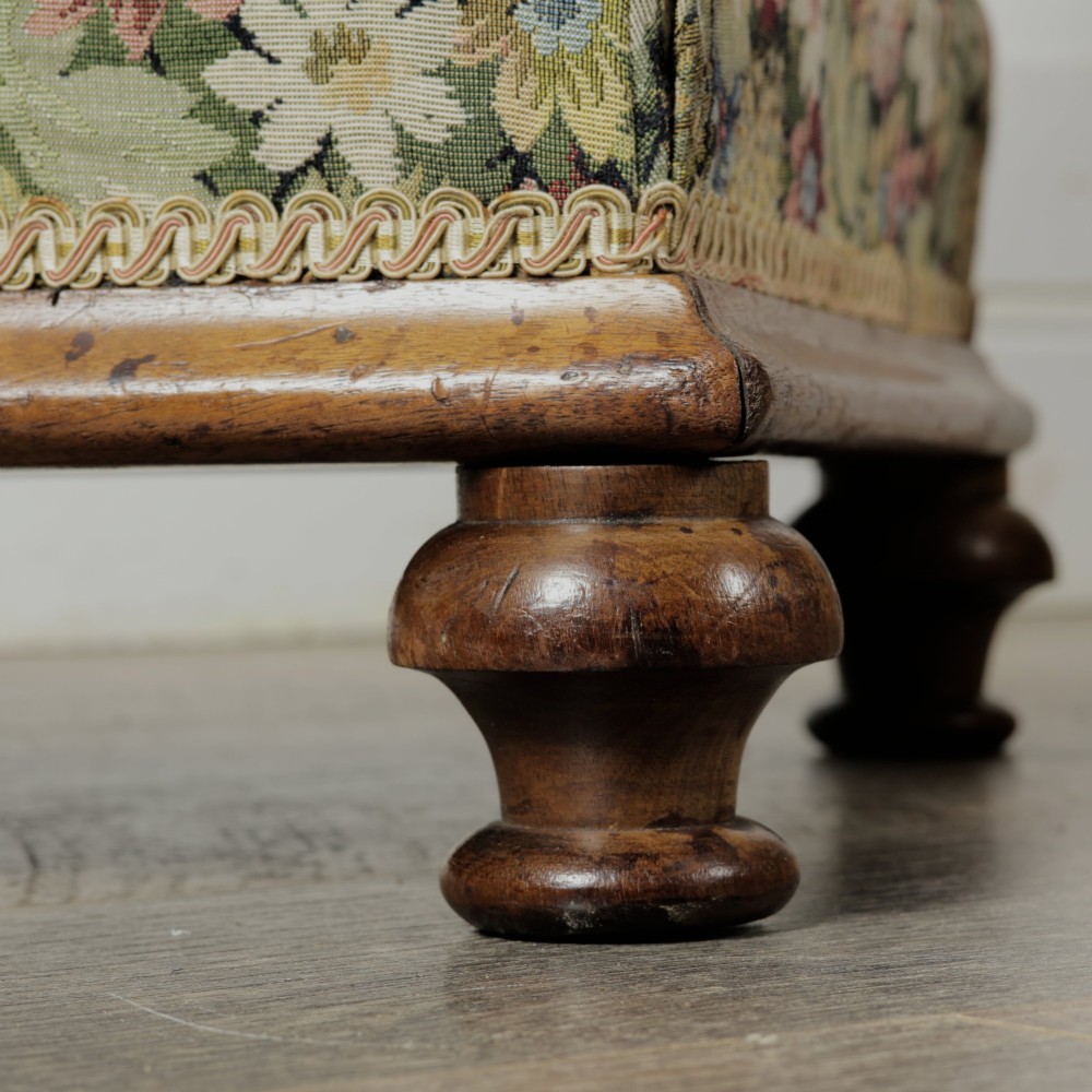 A low Victorian square footstool with green floral tapestry upholstery, decorative braid trim, and turned mahogany feet. View VI