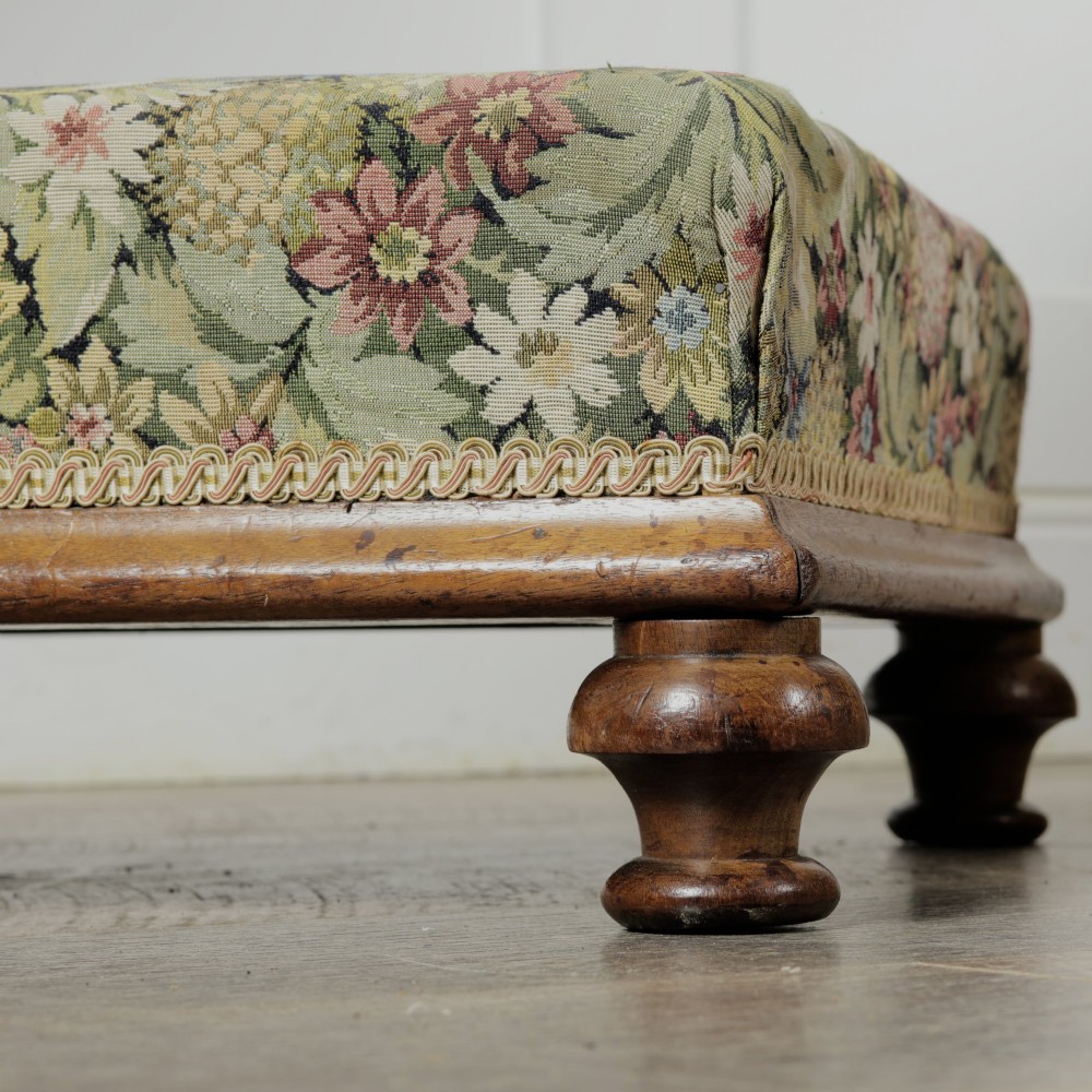 A low Victorian square footstool with green floral tapestry upholstery, decorative braid trim, and turned mahogany feet. View VII