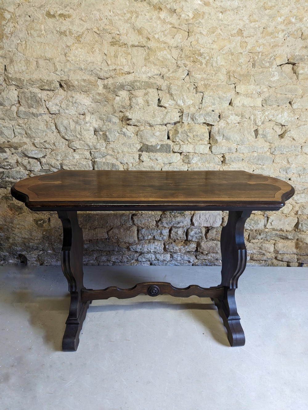 A late 19th-century dark oak trestle table featuring a shaped top with contrasting inlaid border and a low stretcher with a central carved boss.