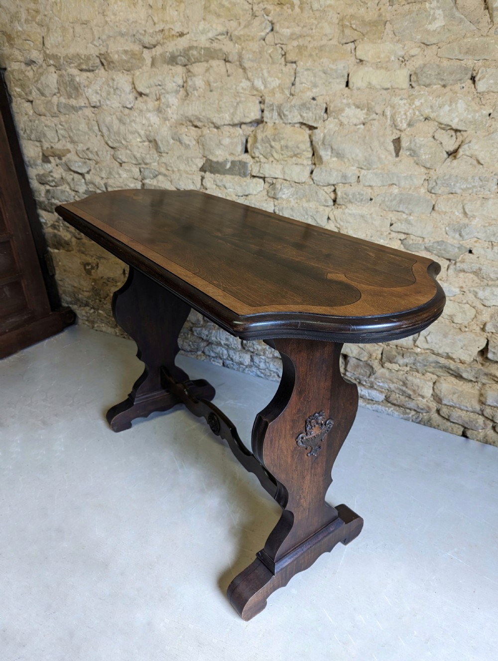A late 19th-century dark oak trestle table featuring a shaped top with contrasting inlaid border and a low stretcher with a central carved boss. View II