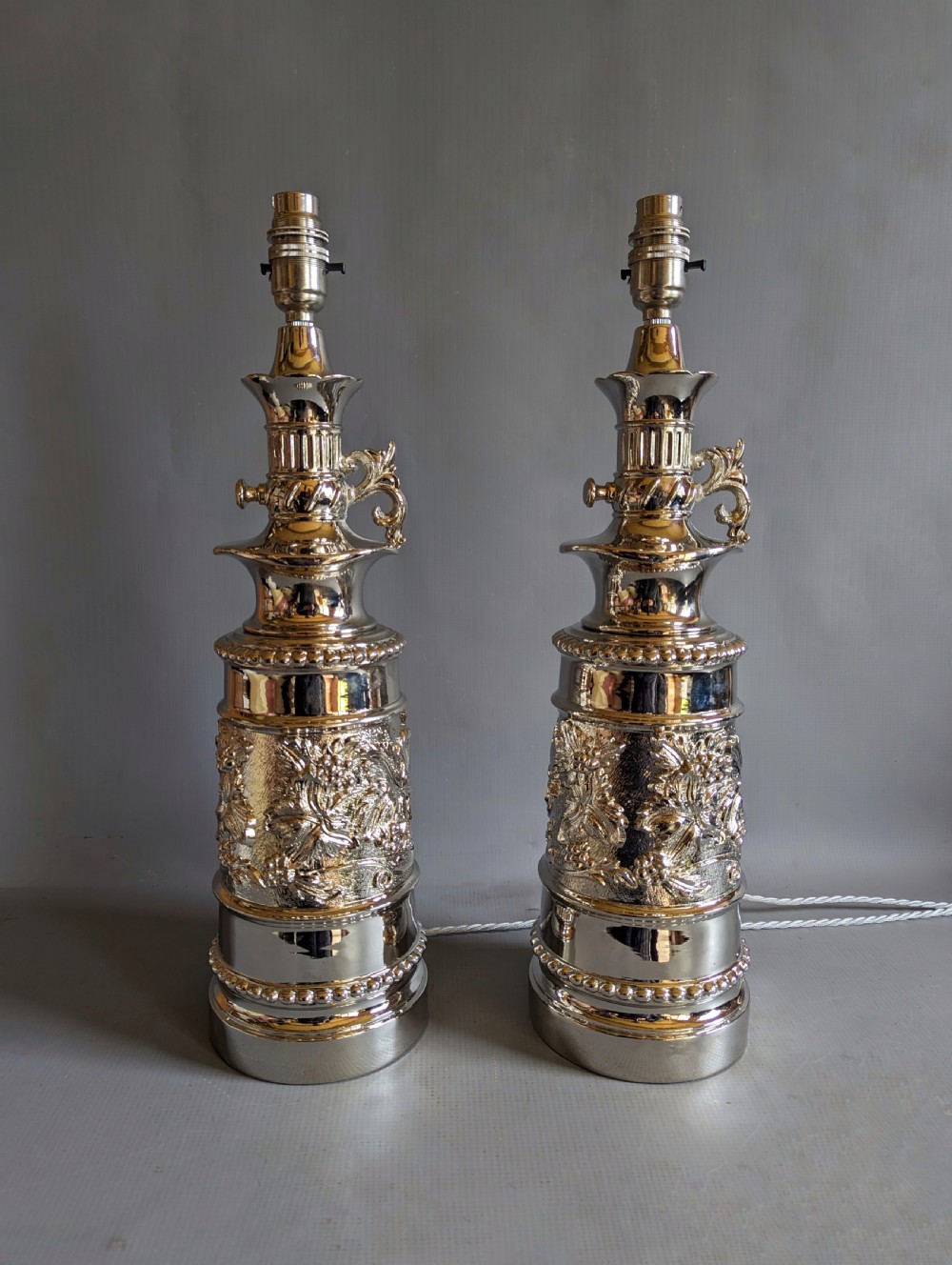A pair of reflective nickel-plated table lamps featuring embossed foliate relief, scroll handles, and beaded rim details.