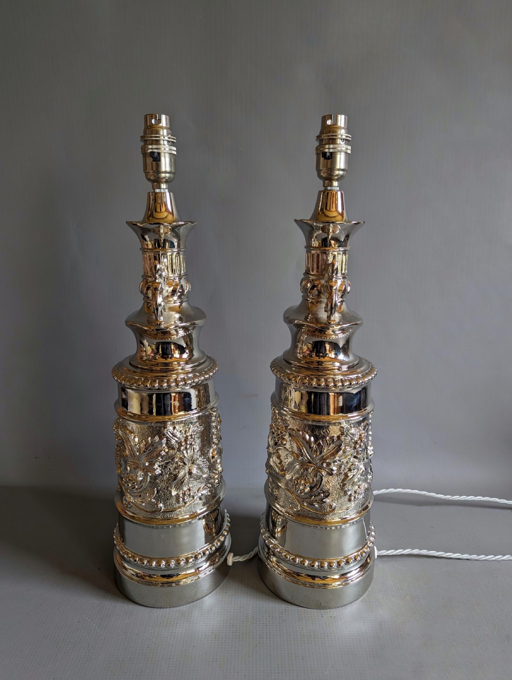 A pair of reflective nickel-plated table lamps featuring embossed foliate relief, scroll handles, and beaded rim details. View IV