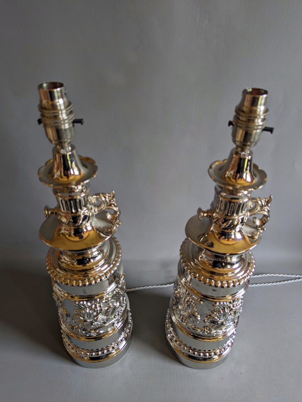 A pair of reflective nickel-plated table lamps featuring embossed foliate relief, scroll handles, and beaded rim details. View V