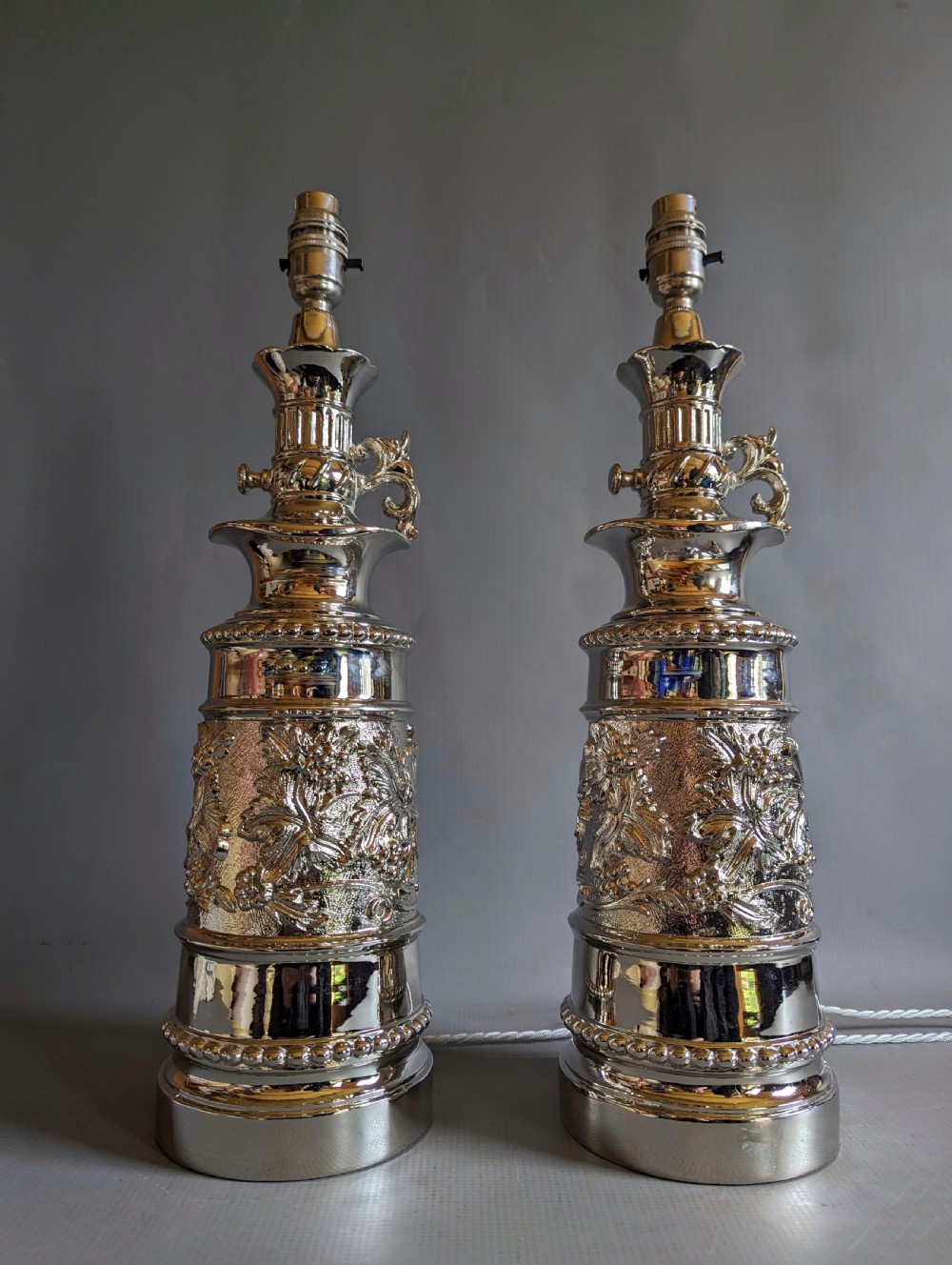 A pair of reflective nickel-plated table lamps featuring embossed foliate relief, scroll handles, and beaded rim details. View VII